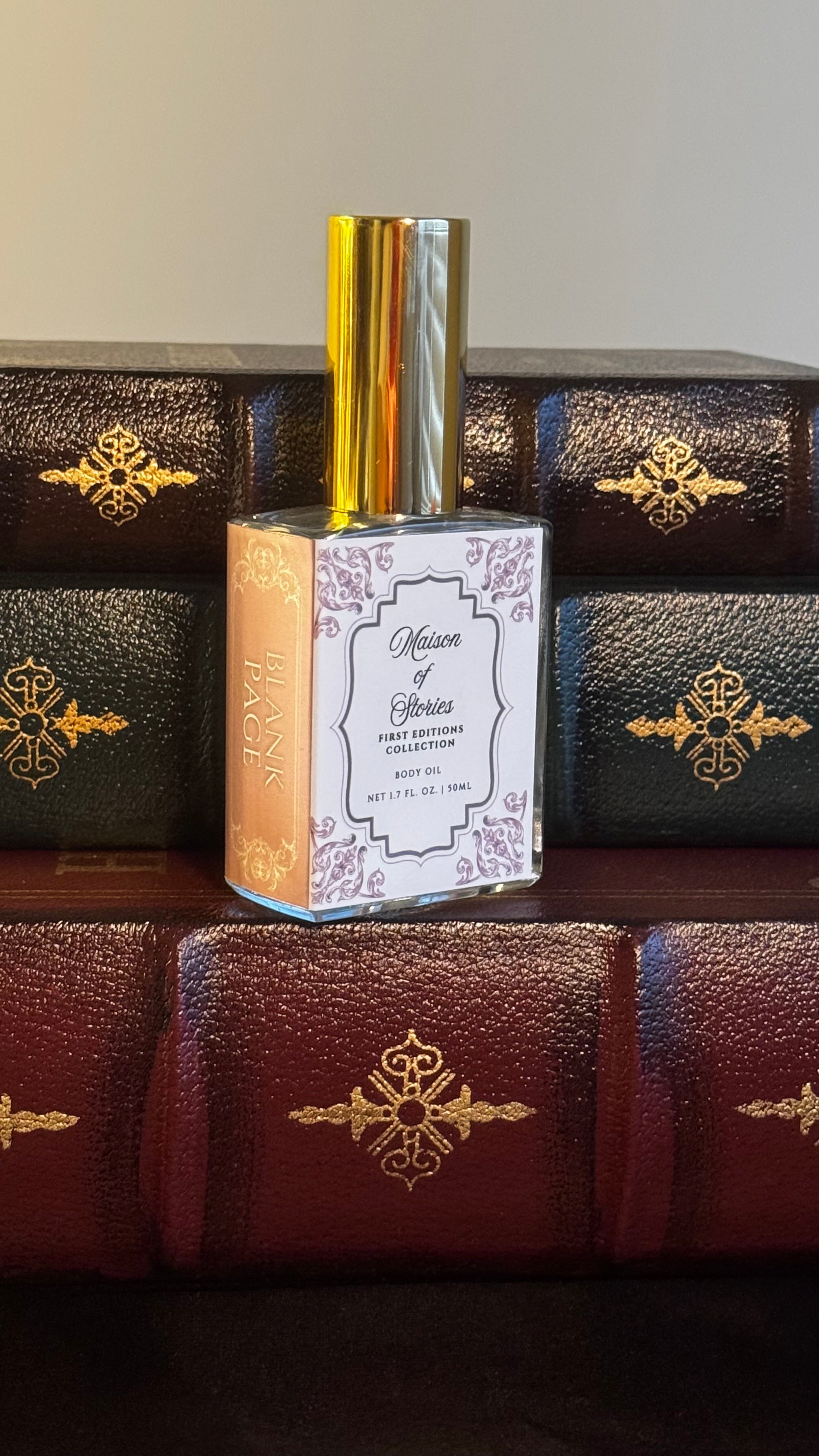 Maison of Stories Blank Page body oil 50ml bottle with rococo floral label and gold cap, displayed on a stack of vintage leather-bound books
