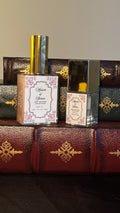 Maison of Stories First Editions Collection body oil size comparison showing 50ml full size and 15ml bookmark size bottles with rococo floral labels on vintage leather-bound books
