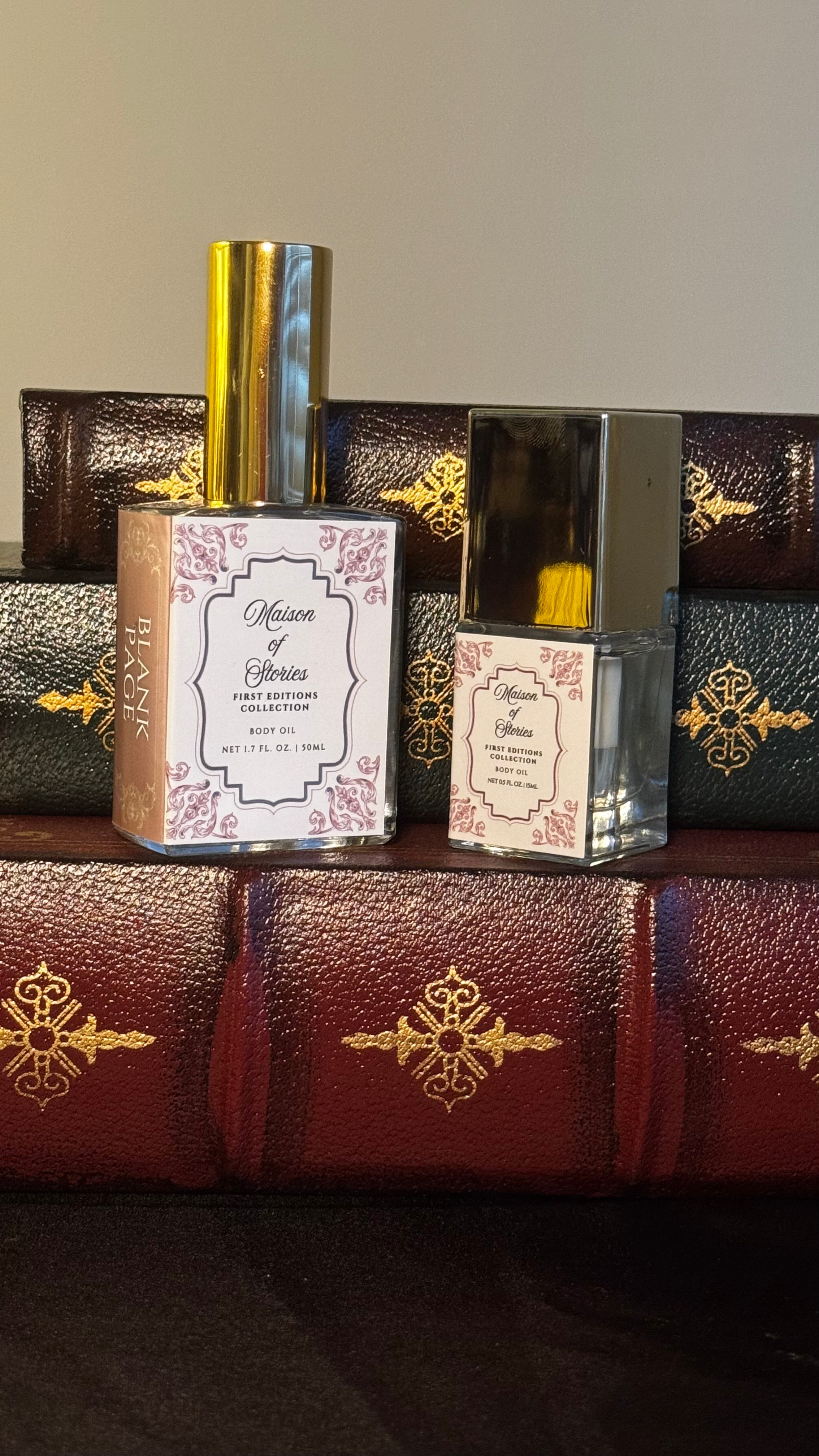 Maison of Stories First Editions Collection body oil size comparison showing 50ml full size and 15ml bookmark size bottles with rococo floral labels on vintage leather-bound books