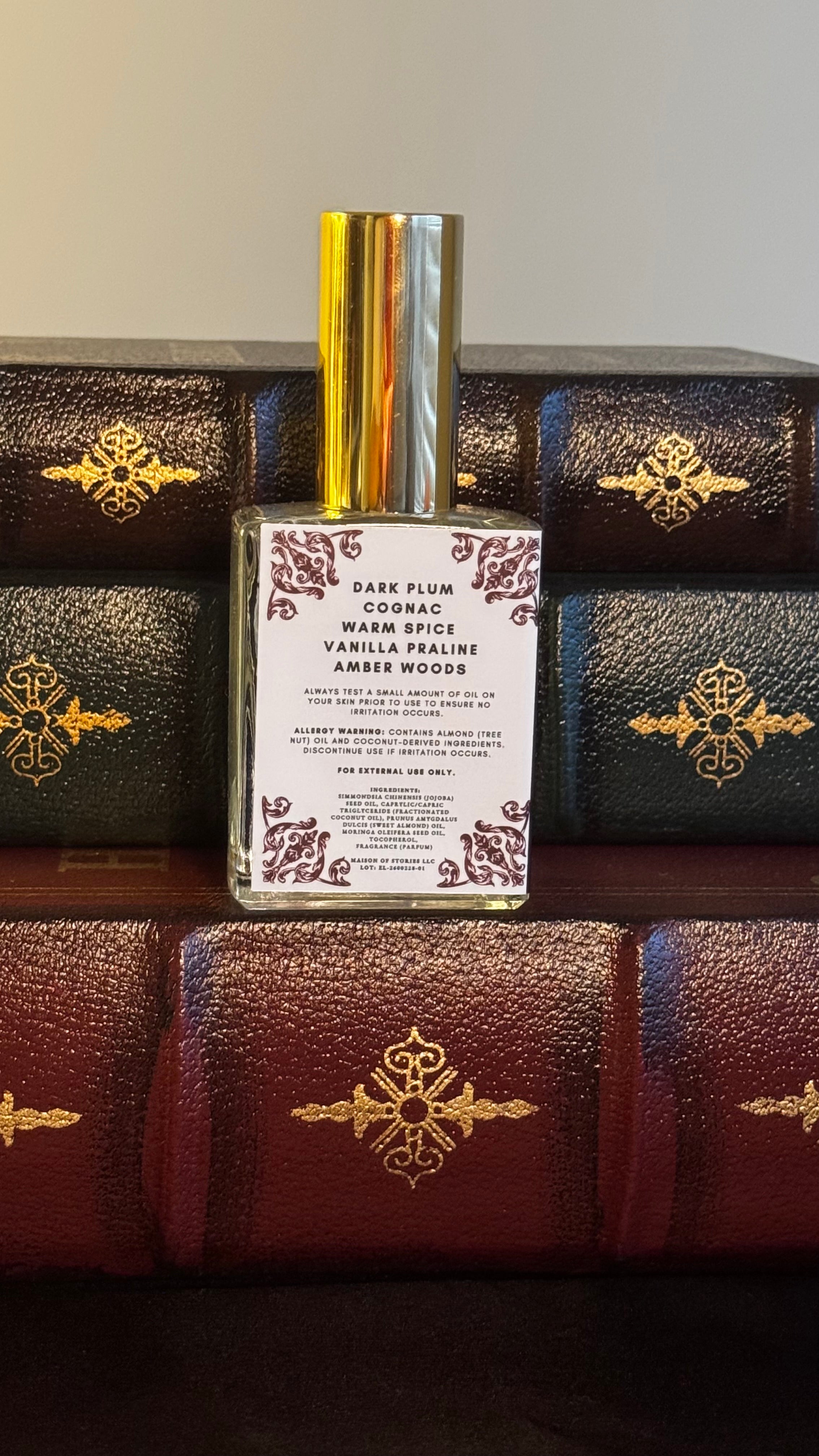 Back label of Maison of Stories Enemies to Lovers body oil 50ml with rococo floral corner details and dark plum, cognac, warm spice, vanilla praline and amber woods scent notes, displayed on vintage leather-bound books

