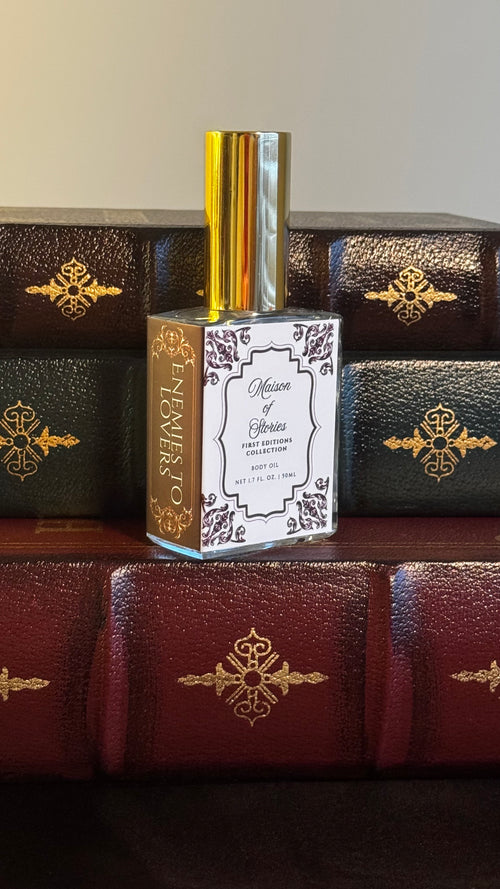 Maison of Stories Enemies to Lovers body oil 50ml bottle with rococo floral label and gold cap, displayed on a stack of vintage leather-bound books