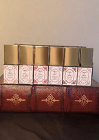 Maison of Stories First Editions Collection six body oil 15ml bottles with rococo floral labels and gold caps displayed on vintage leather-bound books