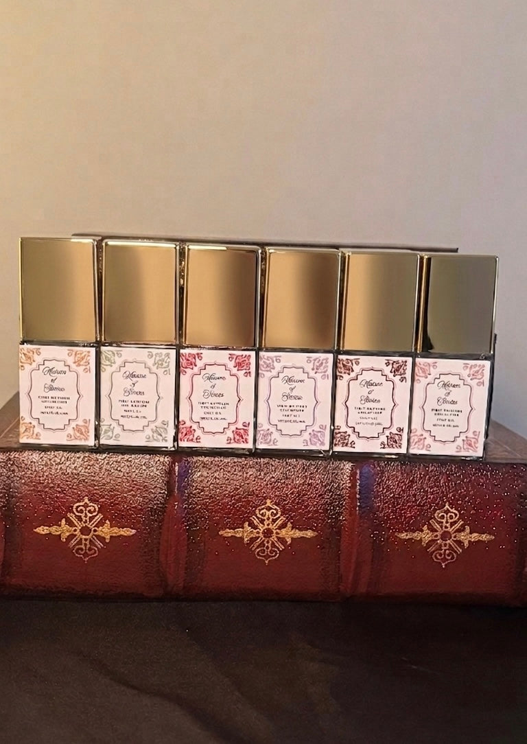 Maison of Stories First Editions Collection six body oil 15ml bottles with rococo floral labels and gold caps displayed on vintage leather-bound books