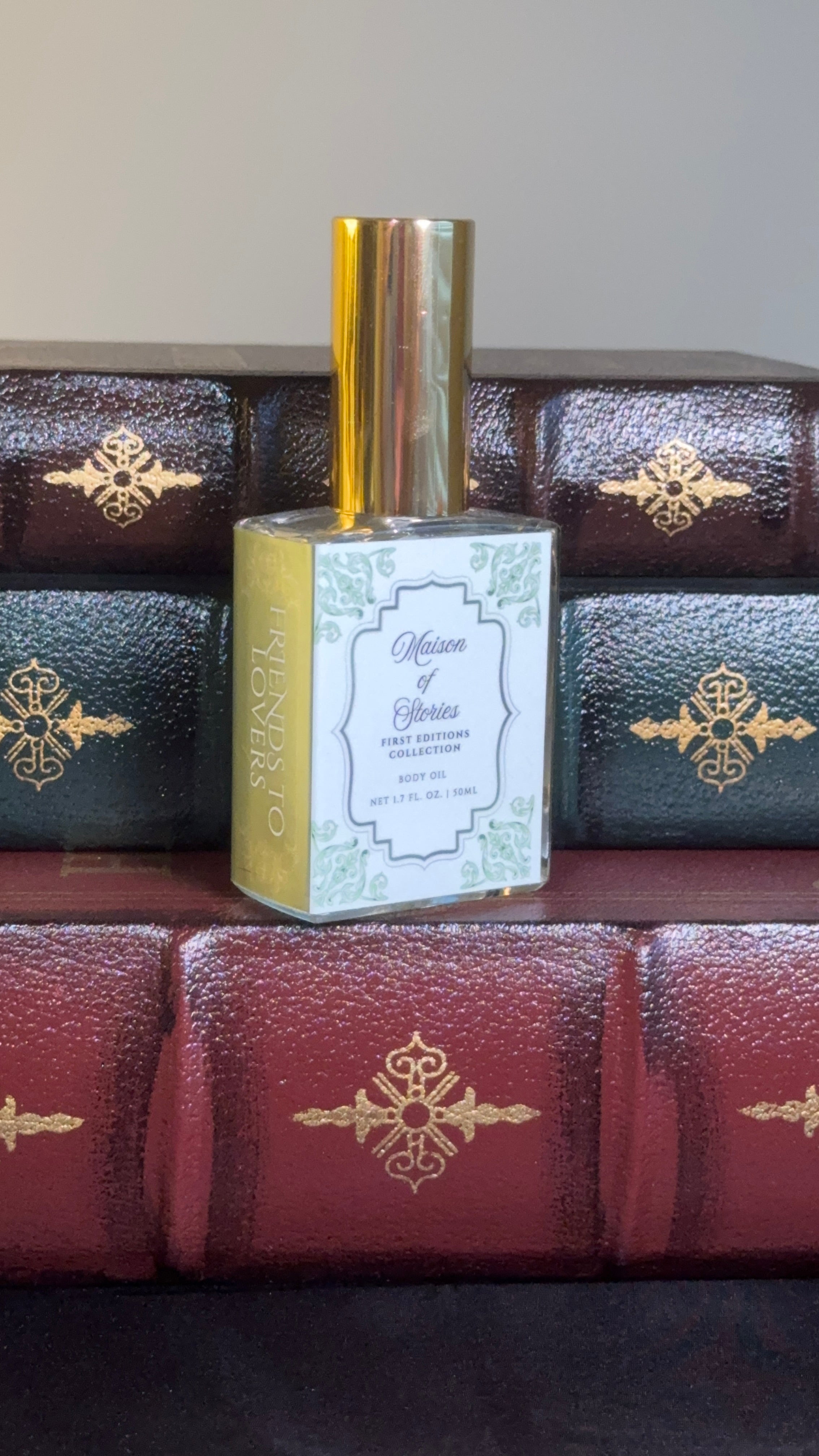 Maison of Stories Friends to Lovers body oil 50ml bottle with rococo floral label and gold cap, displayed on a stack of vintage leather-bound books