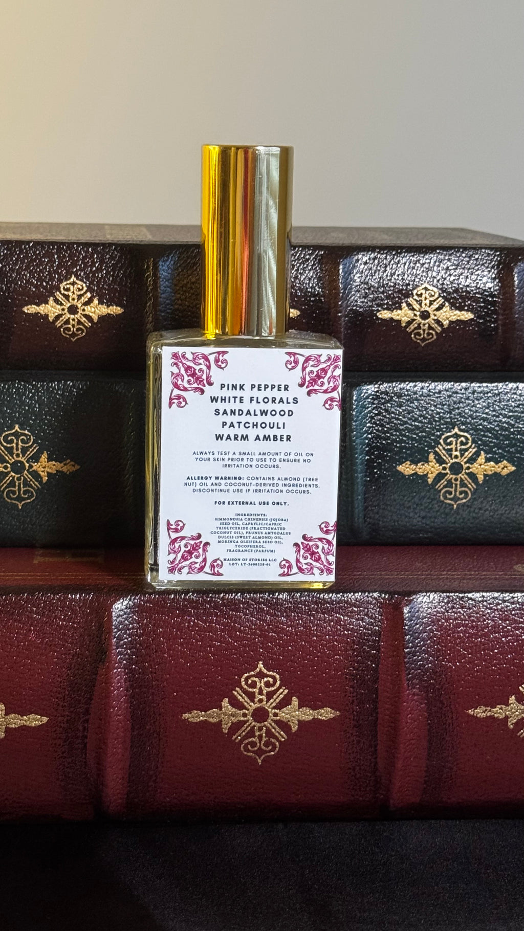 Back label of Maison of Stories Love Triangle body oil 50ml with rococo floral corner details and pink pepper, white florals, sandalwood, patchouli and warm amber scent notes, displayed on vintage leather-bound books
