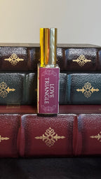 Maison of Stories Love Triangle body oil 50ml spine view with deep magenta label showing scent name, displayed on vintage leather-bound books