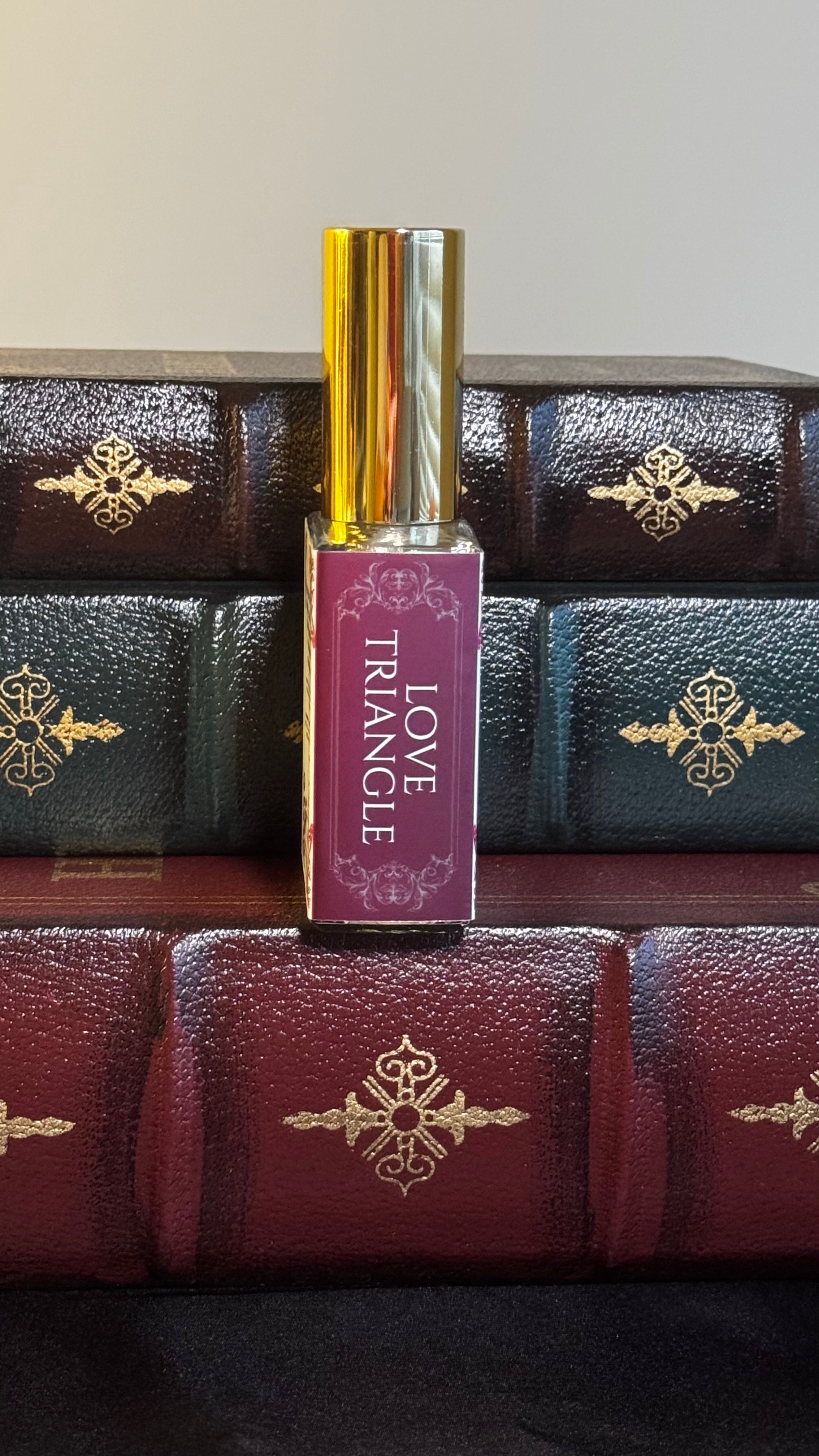 Maison of Stories Love Triangle body oil 50ml spine view with deep magenta label showing scent name, displayed on vintage leather-bound books