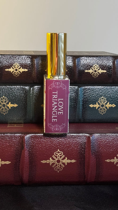 Maison of Stories Love Triangle body oil 50ml spine view with deep magenta label showing scent name, displayed on vintage leather-bound books