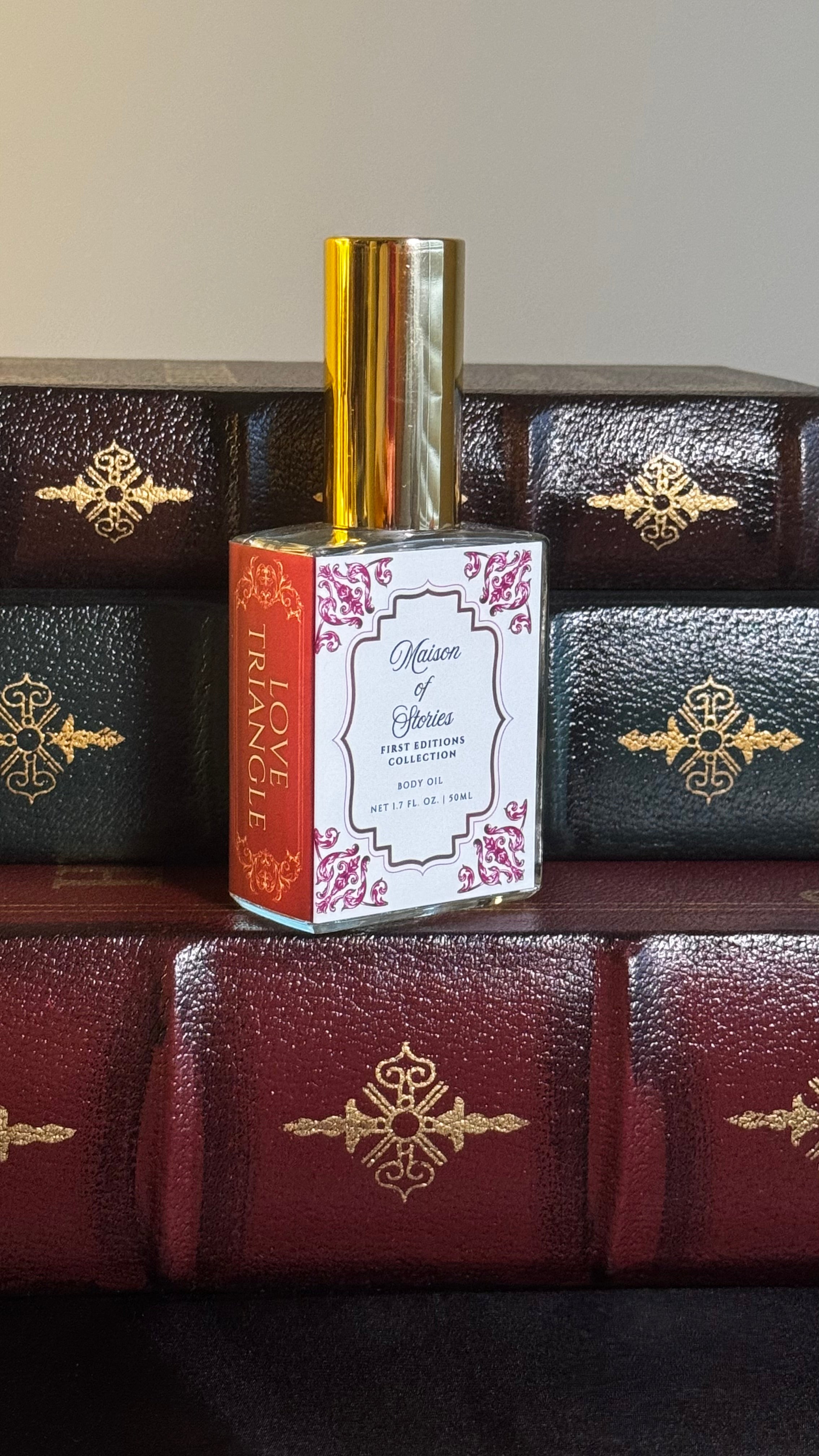 Maison of Stories Love Triangle body oil 50ml bottle with rococo floral label and gold cap, displayed on a stack of vintage leather-bound books