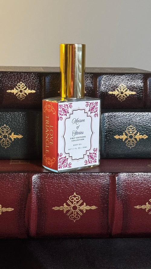 Maison of Stories Love Triangle body oil 50ml bottle with rococo floral label and gold cap, displayed on a stack of vintage leather-bound books