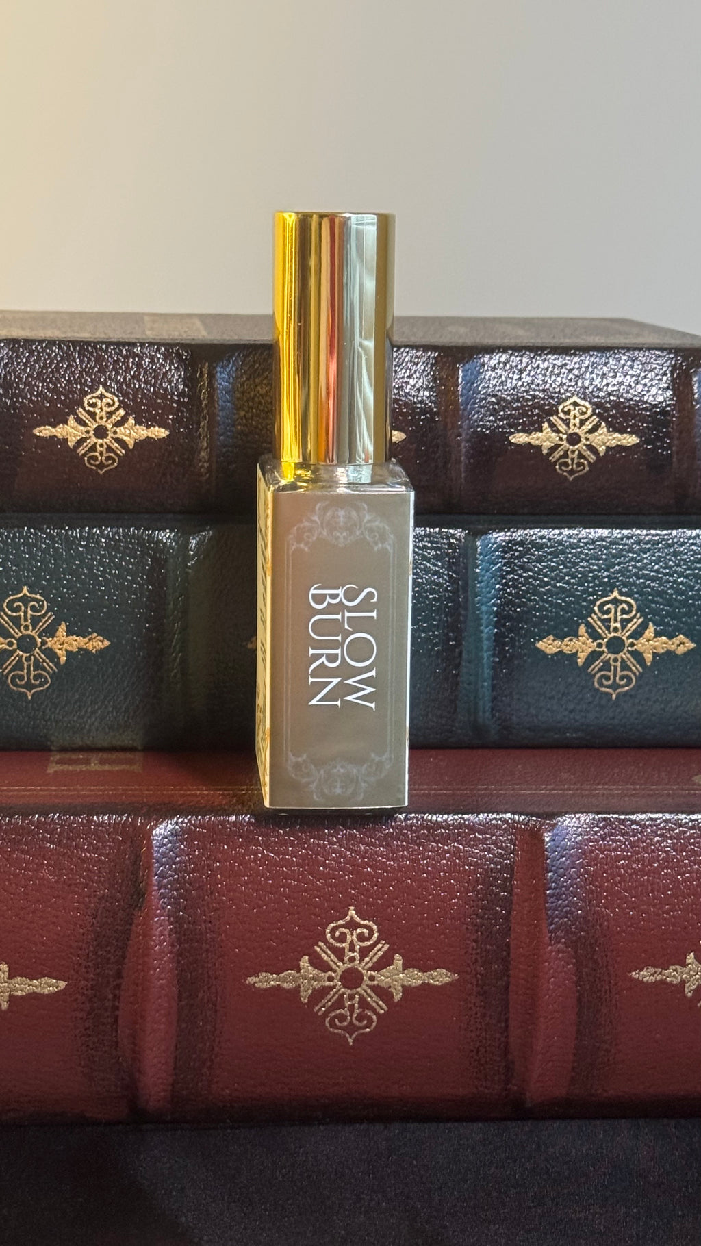 Maison of Stories Slow Burn body oil 50ml spine view with bronzy gold label showing scent name, displayed on vintage leather-bound books