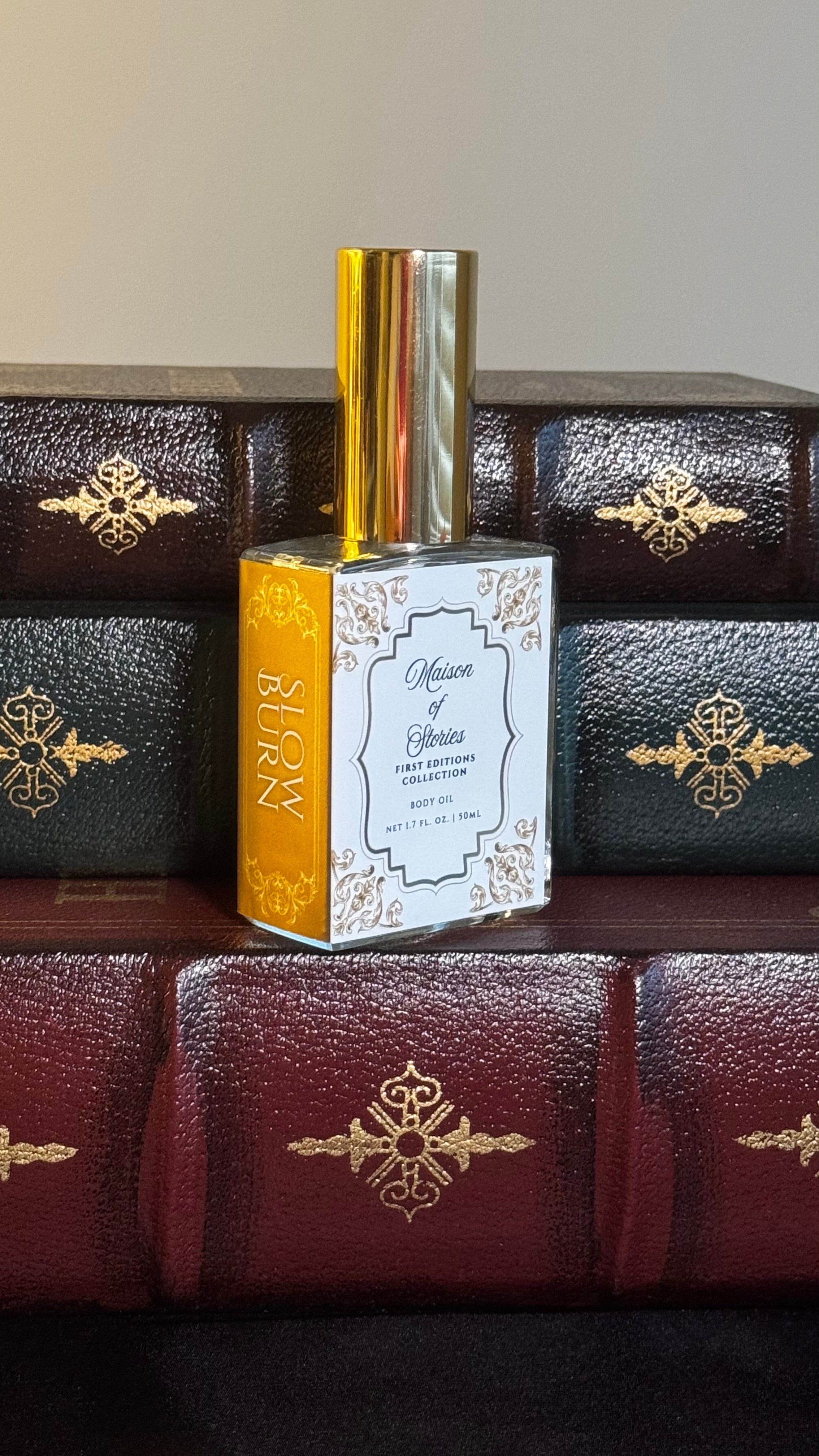 Maison of Stories Slow Burn body oil 50ml bottle with rococo floral label and gold cap, displayed on a stack of vintage leather-bound books