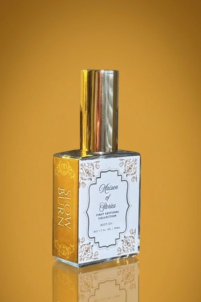 Maison of Stories Slow Burn body oil bottle with gold filigree label and gold cap against a warm amber background